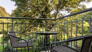 Ein Balkon mit zwei Korbstühlen und einem kleinen runden Tisch, umgeben von einem Metallgeländer, mit Blick auf einen üppigen Grün Garten.