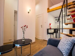 Een moderne woonkamer met een bank, een klein rond tafeltje met een bloevas, een trap met houten leuning, en een keuken in de achtergrond.