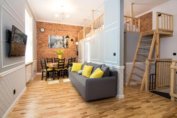 A living room with a grey sofa, wooden floor, brick wall, and a loft area with a wooden ladder. - Apartments Zwierzyniecka A4You (Kraków)