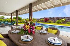 An outdoor dining area with wicker furniture, a pool, and a view of a landscaped garden and mountains.