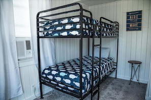 Una cama litera con colcha de rayas azules y blancas de ballenas, una pequeña mesa redonda y un letrero que dice 'RIDE THE WAVE'.