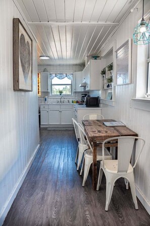 A narrow hallway leading to a kitchen with white cabinets, a wooden table, and white chairs.