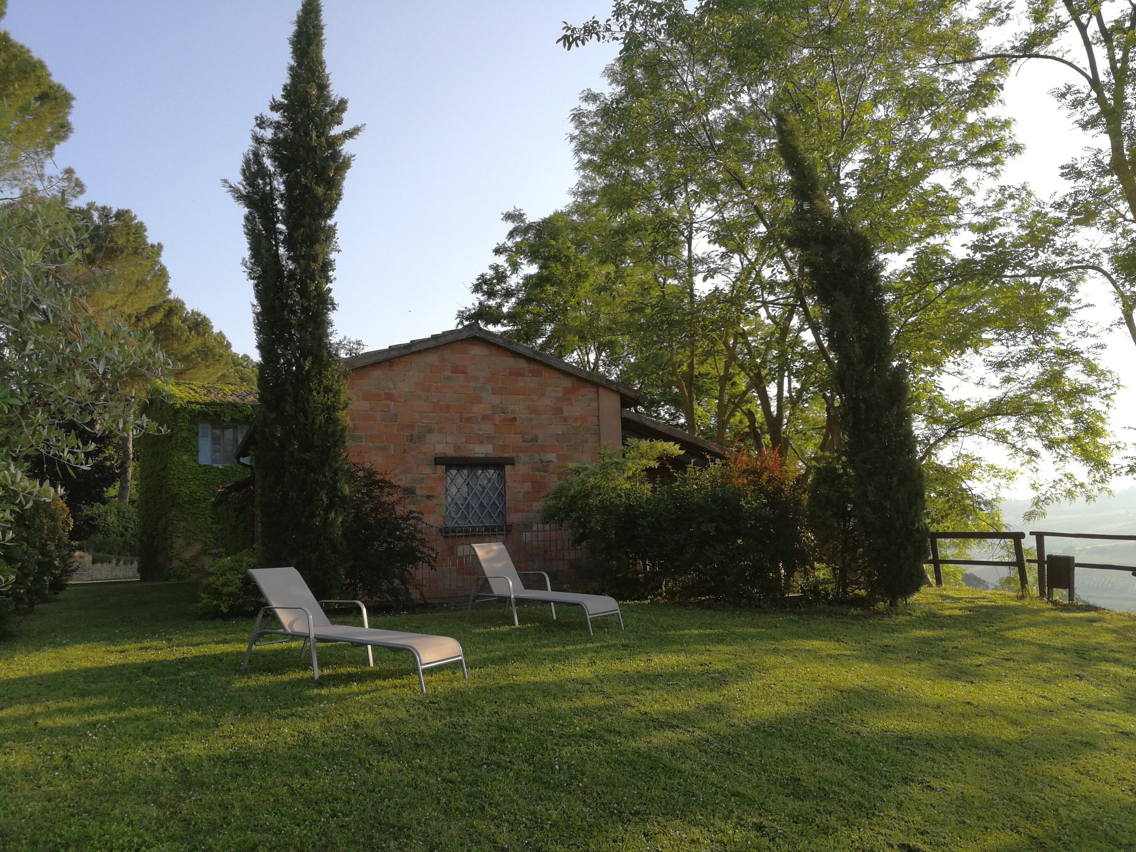 A brick building with a window and a door, surrounded by greenery and two white lounge chairs.