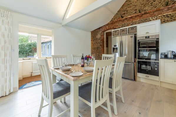 A dining area with a wooden table set for four, white chairs, a brick wall, and a kitchen with modern appliances.
