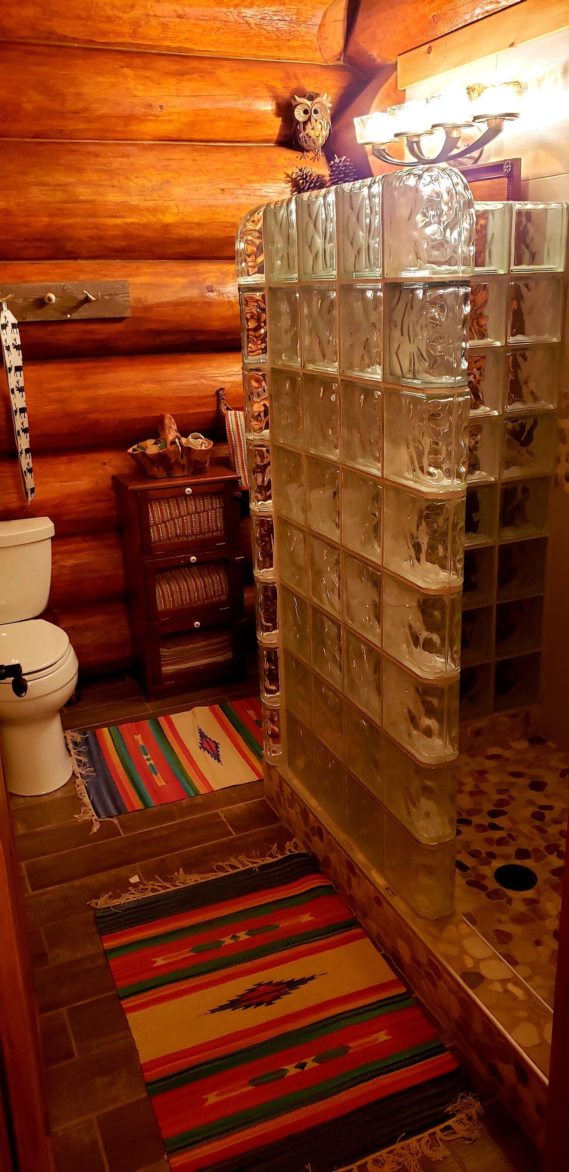 A bathroom with a glass shower enclosure, wooden walls, a white toilet, a small wooden cabinet, and a colorful rug.