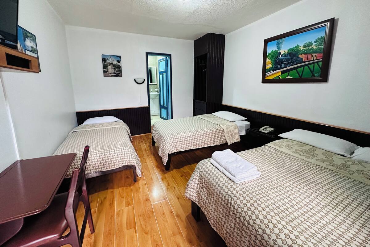 A hotel room with two single beds, a wooden floor, a desk, and a chair. There is a painting on the wall and a door leading to a bathroom.