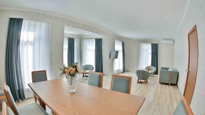 A modern hotel room with a wooden dining table, grey upholstered chairs, a flat-screen TV mounted on the wall, and a sofa. - Yurd Hotel (?lisu)
