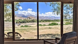 Quarto com uma grande janela com vista para uma paisagem desértica e montanhas. Há duas cadeiras e uma mesa no quarto.