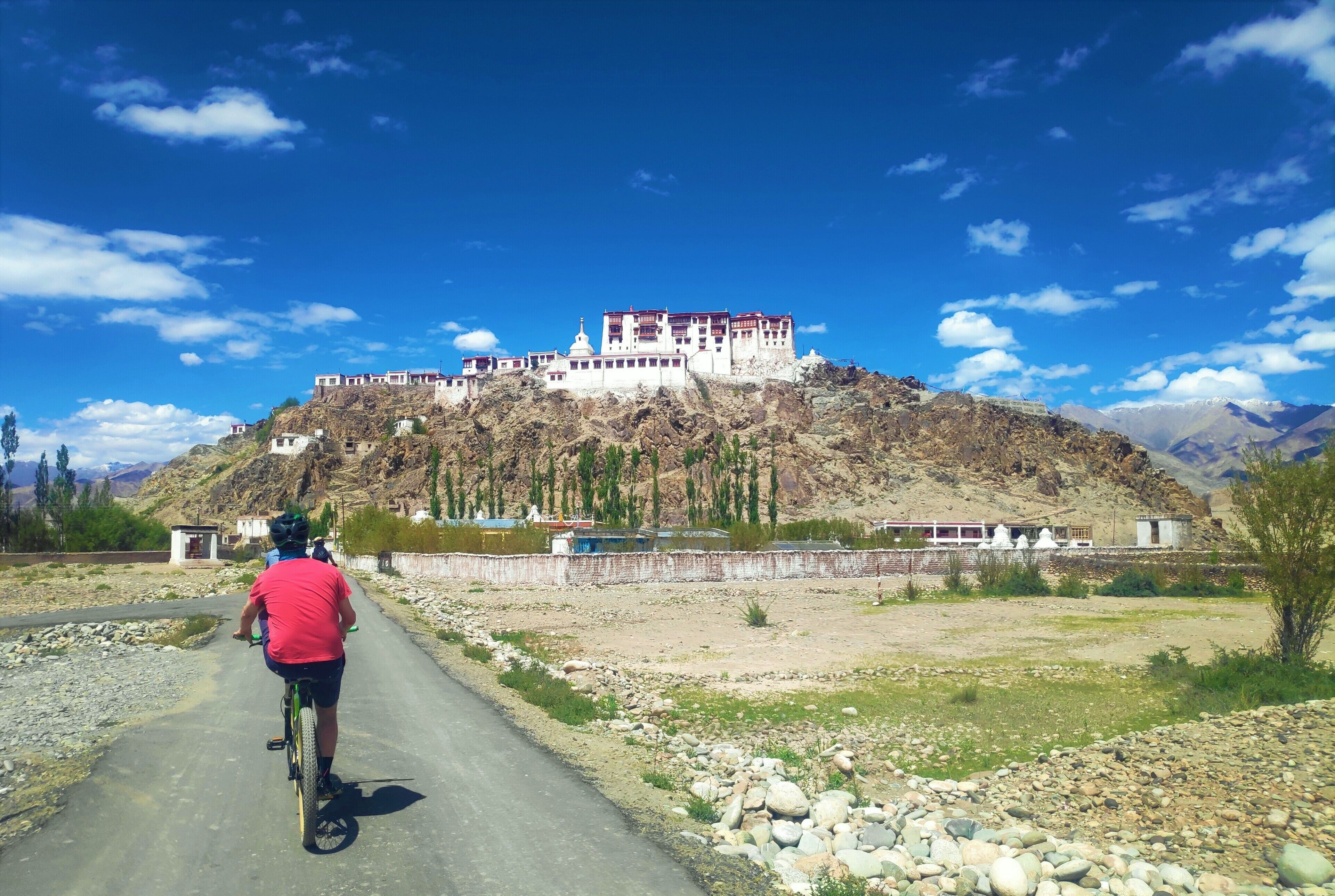 A person riding a bicycle on a paved road with a large, multi-story building on a hill in the background.