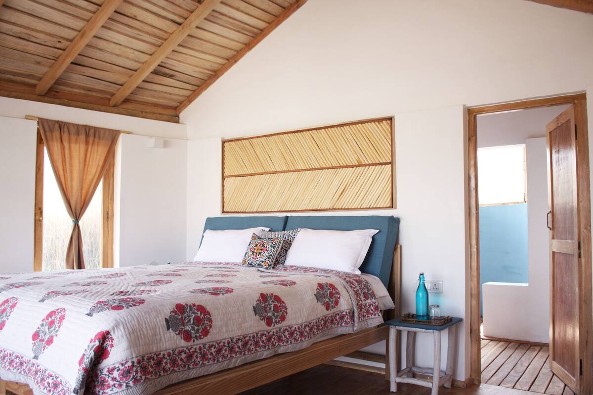 A bedroom with a wooden ceiling, a bed with a floral bedspread, a bedside table with a bottle, and a view of the outside.