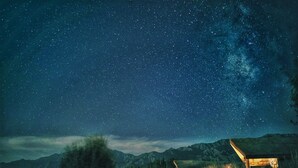 Céu noturno com estrelas e a Via Láctea sobre uma paisagem montanhosa com duas estruturas de madeira.
