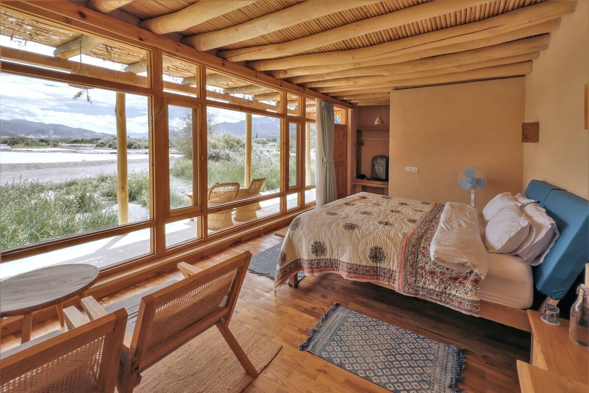 A room with a bed, a table, and chairs. There are large windows with a view of the outdoors.