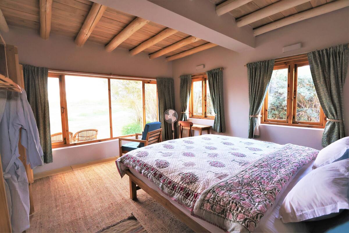 A bedroom with a large bed, wooden ceiling beams, a desk, and a chair near a window.