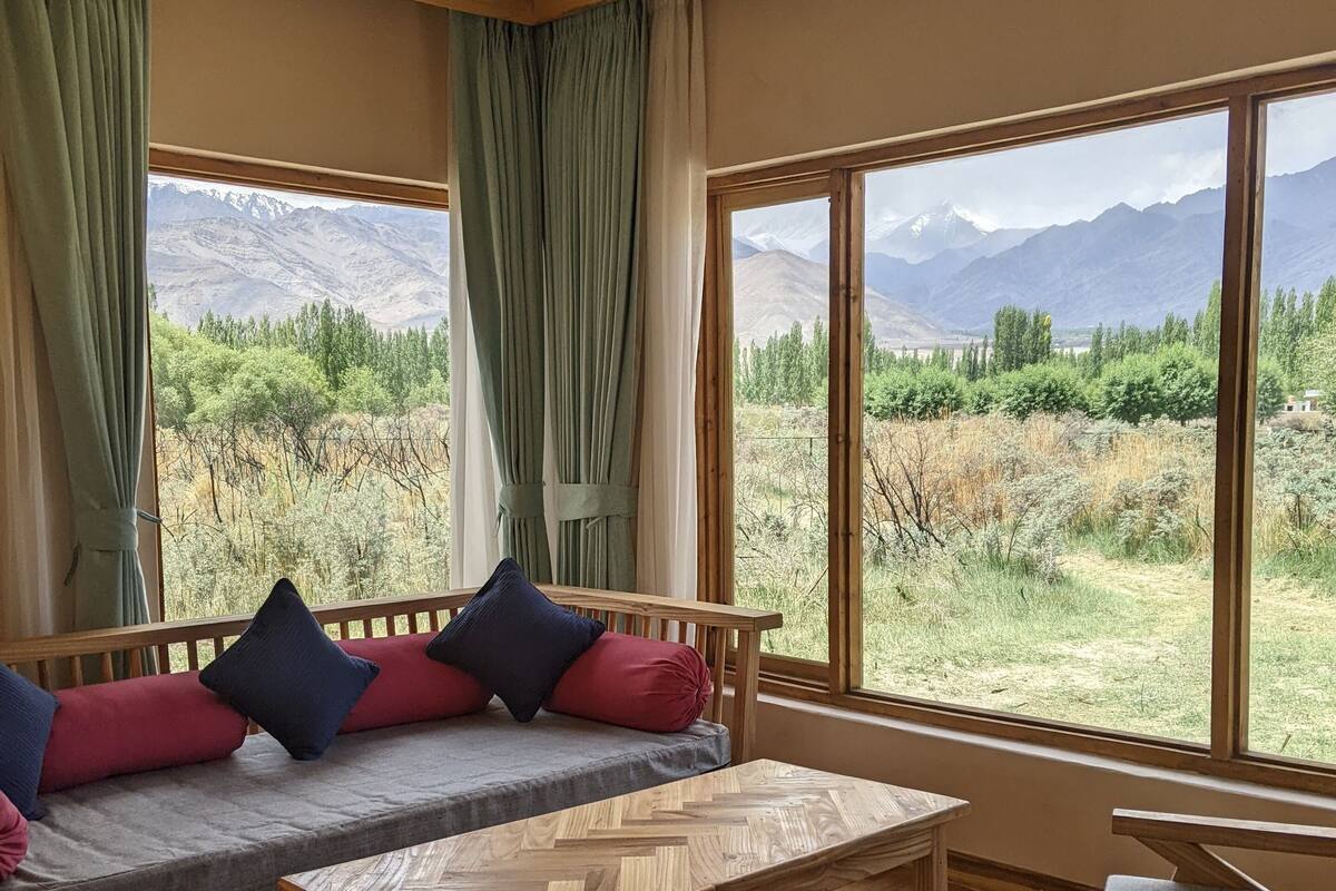 A cozy room with a wooden ceiling, a sofa with cushions, a wooden table, and large windows offering a mountain view.