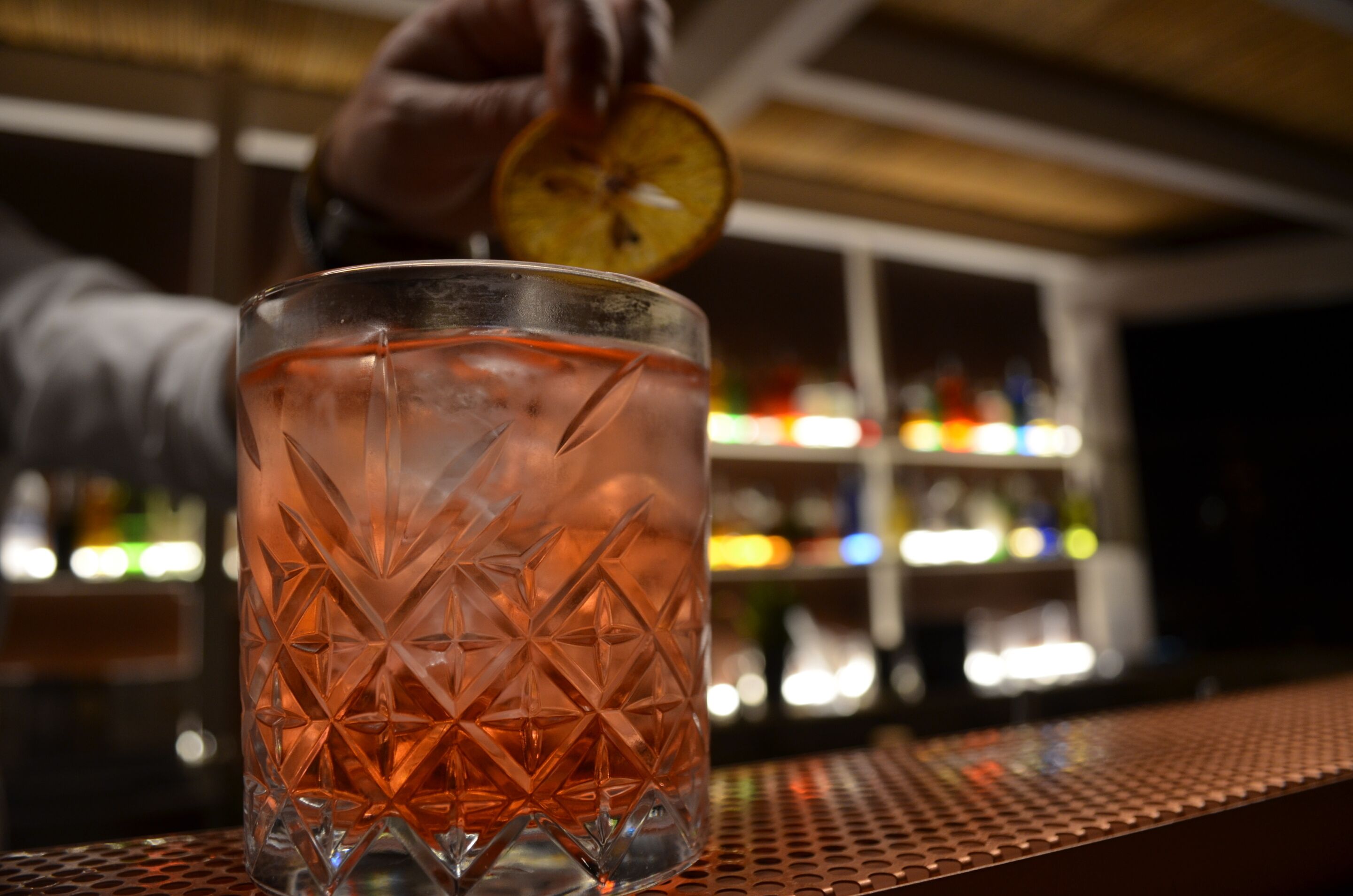 A bar setting with a glass of amber-colored drink, a hand holding a slice of citrus, and a blurred background with shelves of bottles.