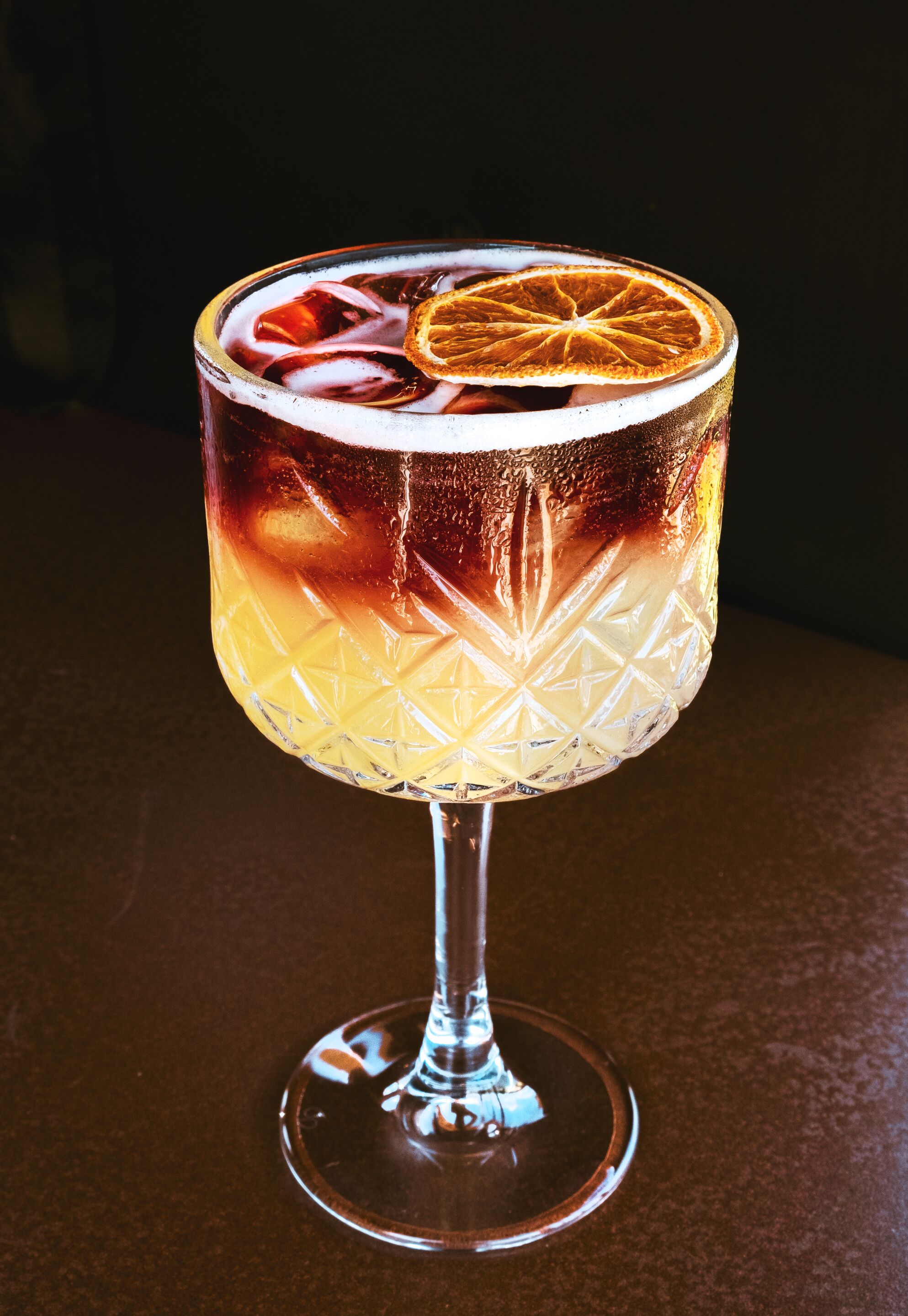 Un bicchiere da cocktail con motivo decorativo, contenente una bevanda scura e una fetta d'arancia.