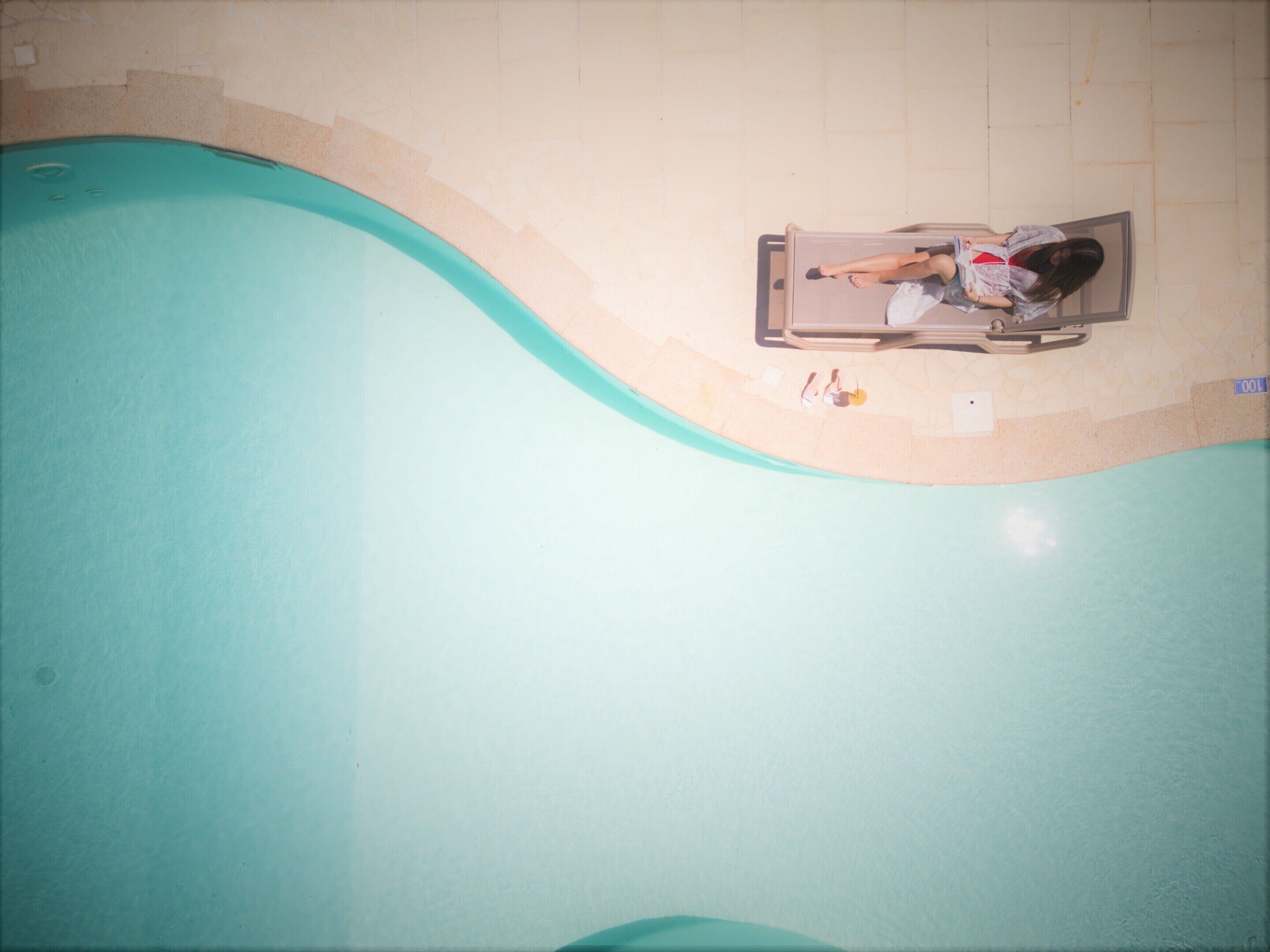 A person lying on a lounge chair by a pool with a tiled floor and a light-colored wall.