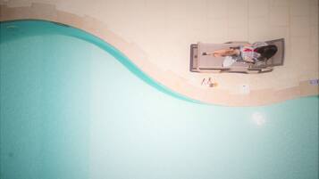 A person lying on a lounge chair by a pool with a tiled floor and a light-colored wall.