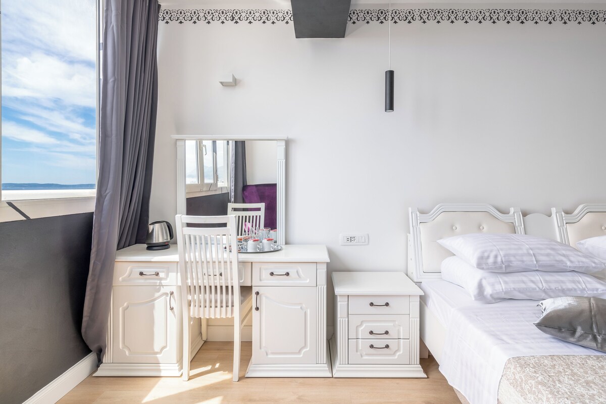 A bedroom with a bed, bedside table, dresser, and a chair. There is a window with curtains, a view of the sky, and a wall with a decorative border.