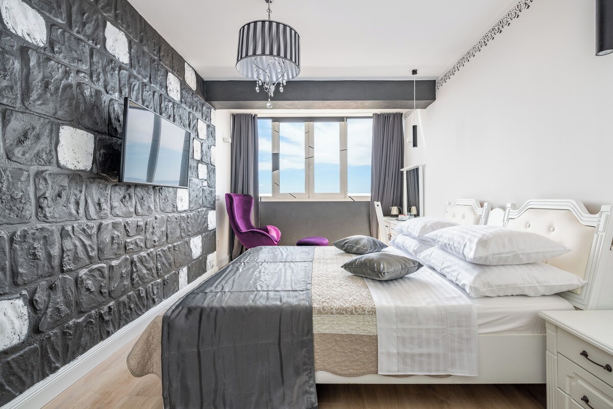 A modern bedroom with a stone wall, a bed with white bedding, a purple chair, and a large window with a view.