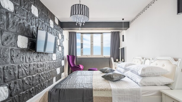 Une chambre moderne avec un mur en pierre, un lit avec une literie blanche, un fauteuil violet et une grande fenêtre donnant sur un beau paysage.