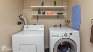 A laundry area with two washing machines, a vent, shelves with plants and a box, and a towel hanging on a hook. - Eagle's Haven - Lower - 2 bedroom, 1 Bath (Cooper Landing)