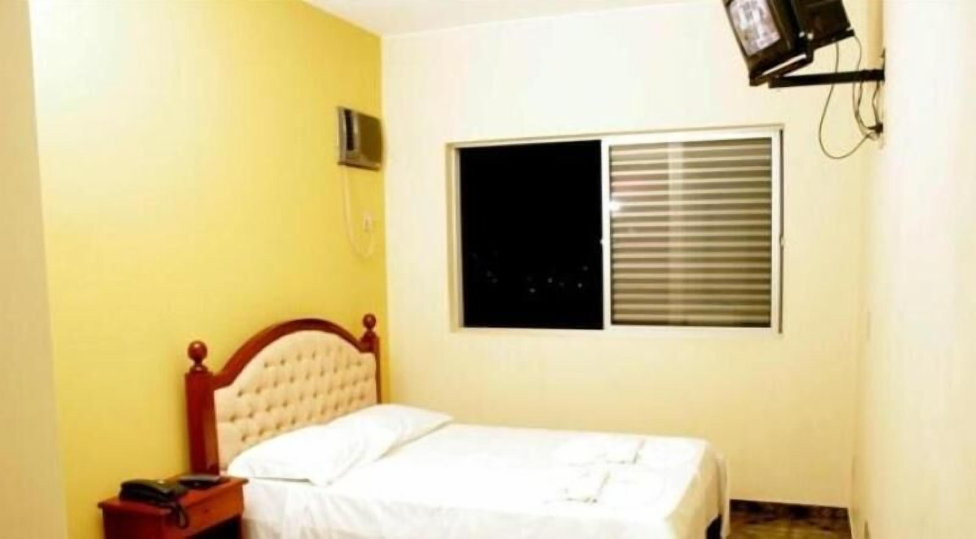 Quarto com cama, mesa de cabeceira, televisão na parede e janela com persianas.