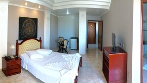 Habitación de hotel con cama, mesita de noche, lámpara, silla, escritorio, televisor y baño visible a través de una puerta abierta.