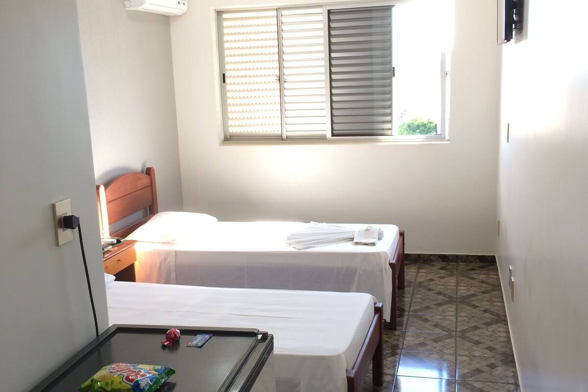 A small, clean room with two beds, a refrigerator, and a window with blinds.