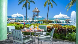 Un espace aménagé au bord de la piscine, avec des chaises longues, une table où sont servis les articles du petit-déjeuner, des palmiers et une vue sur l’océan.
