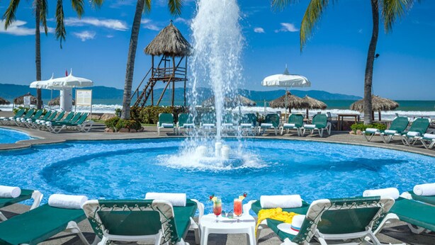 Une piscine dotée d’une fontaine centrale, entourée de chaises longues et de palmiers, et offrant une vue sur l’océan.