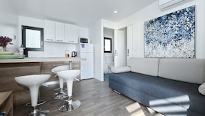 A modern kitchen with white cabinets, a microwave, and a coffee maker. There are two white bar stools and a gray sofa in the living area.