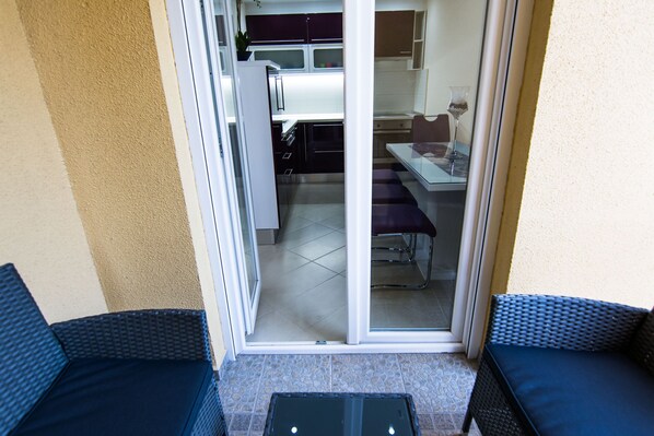 A balcony with two wicker chairs, a small table, and a sliding glass door leading to a kitchen area with a dining table and chairs.