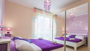 A modern bedroom with a large mirror, a bed with purple bedding, a nightstand with a lamp, and a window with colorful blinds. - Apartment Songbird (Biograd)