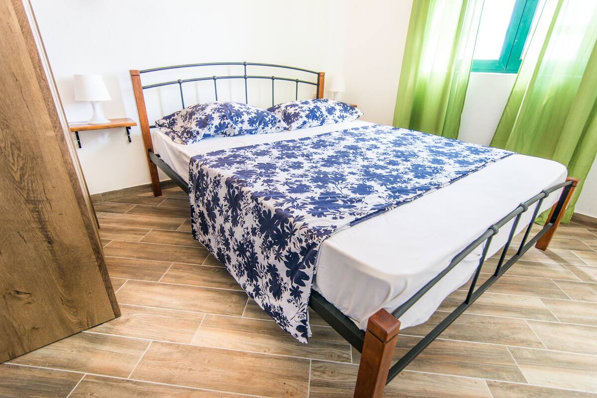 A neatly made bed with a floral bedspread and pillows, a wooden nightstand with a lamp, and a window with green curtains.