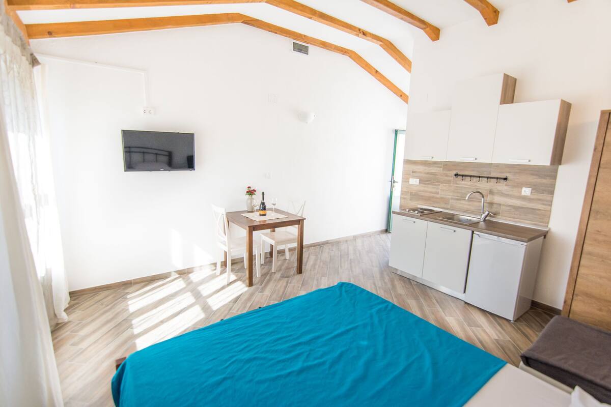 A compact room with a bed, a small table with a chair, a TV mounted on the wall, a kitchenette with cabinets and a sink, and wooden beams on the ceiling.