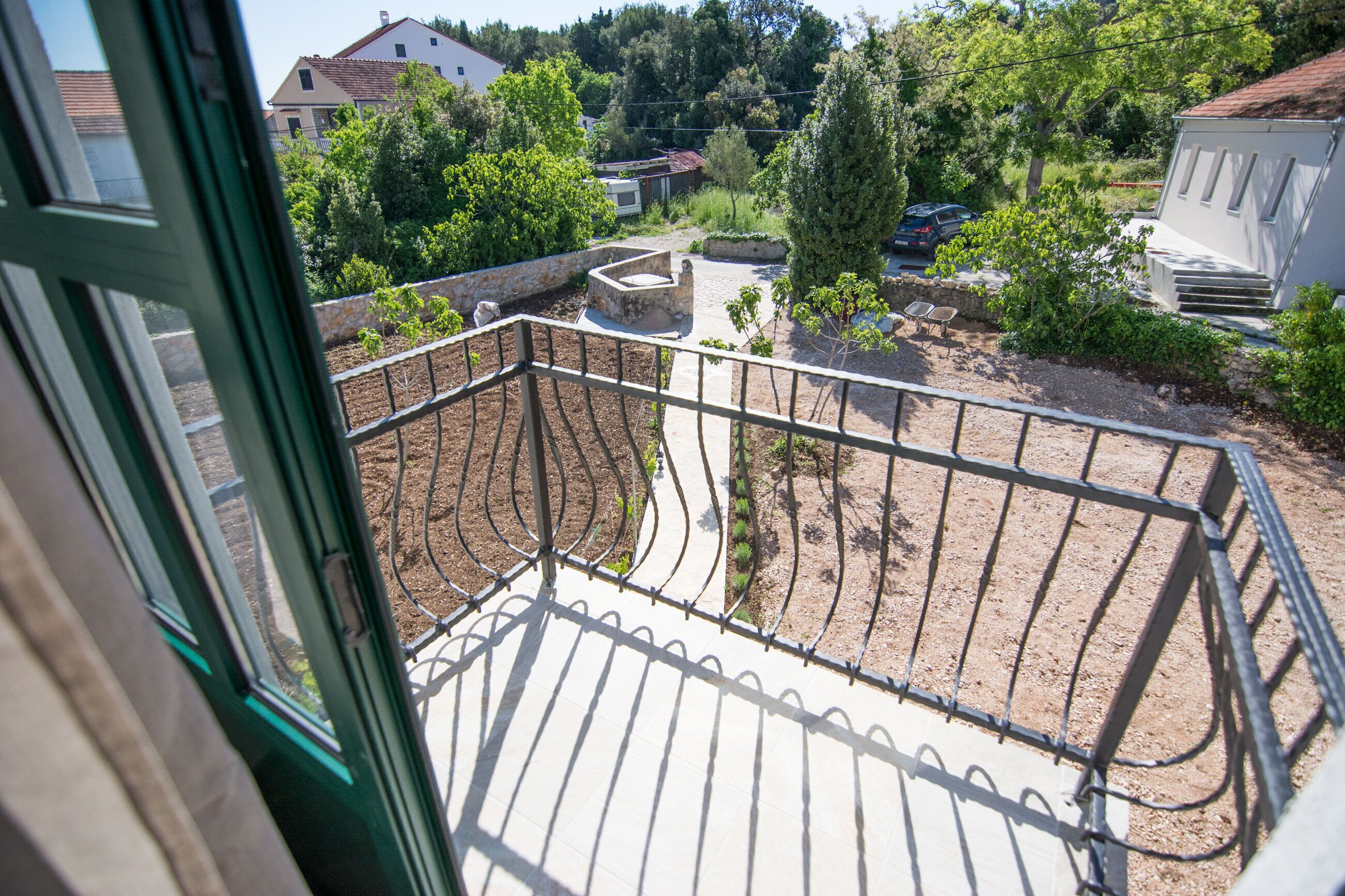 A balcony with a metal railing, a garden area with plants and a small tree, and a view of a residential area with houses and trees.