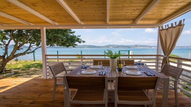 A wooden deck with a table set for two, overlooking a coastal view with a fence and trees. - Mobile Home Frangipani 1 (Biograd)