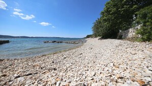 A pebbled beach with clear water, a stone wall, and a large tree. - Mobile Home Frangipani 1 (Biograd)