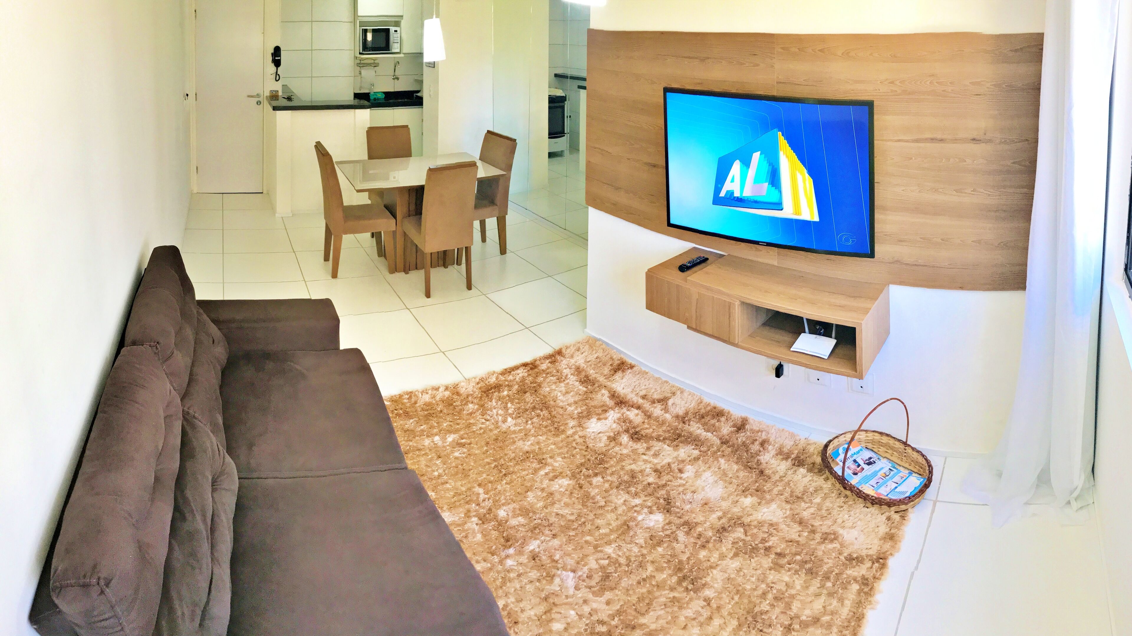 A modern living room with a flat-screen TV mounted on a wooden wall, a brown sofa, a dining area with chairs, and a kitchen in the background.
