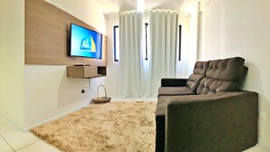 Ein Wohnzimmer mit einem an der Wand montierten Flachbildfernseher, einem braunen Sofa, einem beigefarbenen Teppich und einem Fenster mit weißen Vorhängen.