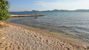 A beach with clear water, a person walking on a pier, and distant mountains.