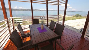 A covered outdoor seating area with a wooden table and chairs, overlooking a body of water and distant hills. - Mobile Home Plumeria 1 (Biograd)