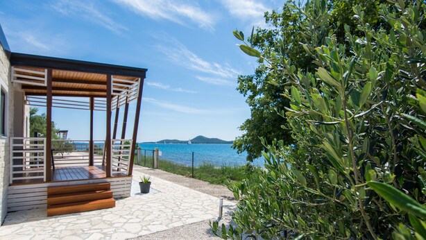 A covered outdoor area with a stone floor leading to a wooden deck, overlooking a body of water and a distant island. - Mobile Home Plumeria 3 (Biograd)