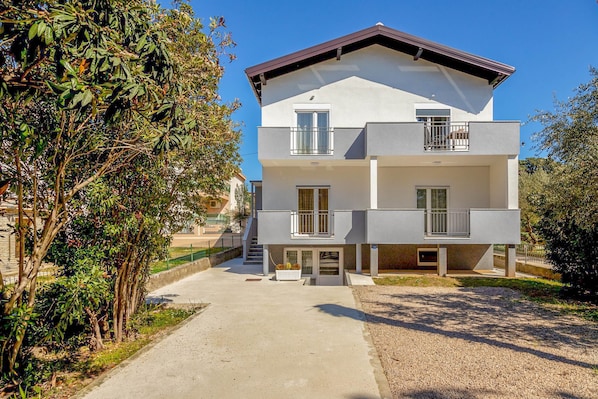 Ein zweigeschossiges Wohnhaus mit einem Balkon im oberen Geschoss, einer Auffahrt zum Eingang und einem Gartenbereich mit Bäumen und Sträuchern.