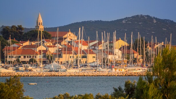 A coastal town with a prominent church tower, numerous sailboats docked along the shore, and a rocky breakwater. - Apartments Grapevines (Biograd)