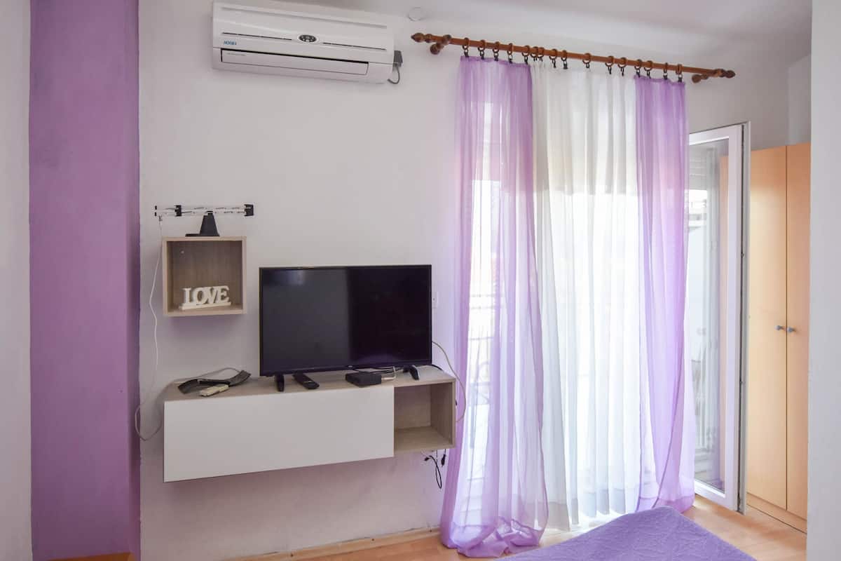 Quarto com televisão em prateleira branca, cortinas roxas e porta de madeira.