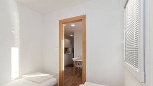 A small, bright room with two beds, a wooden floor, and a door leading to a kitchen area.