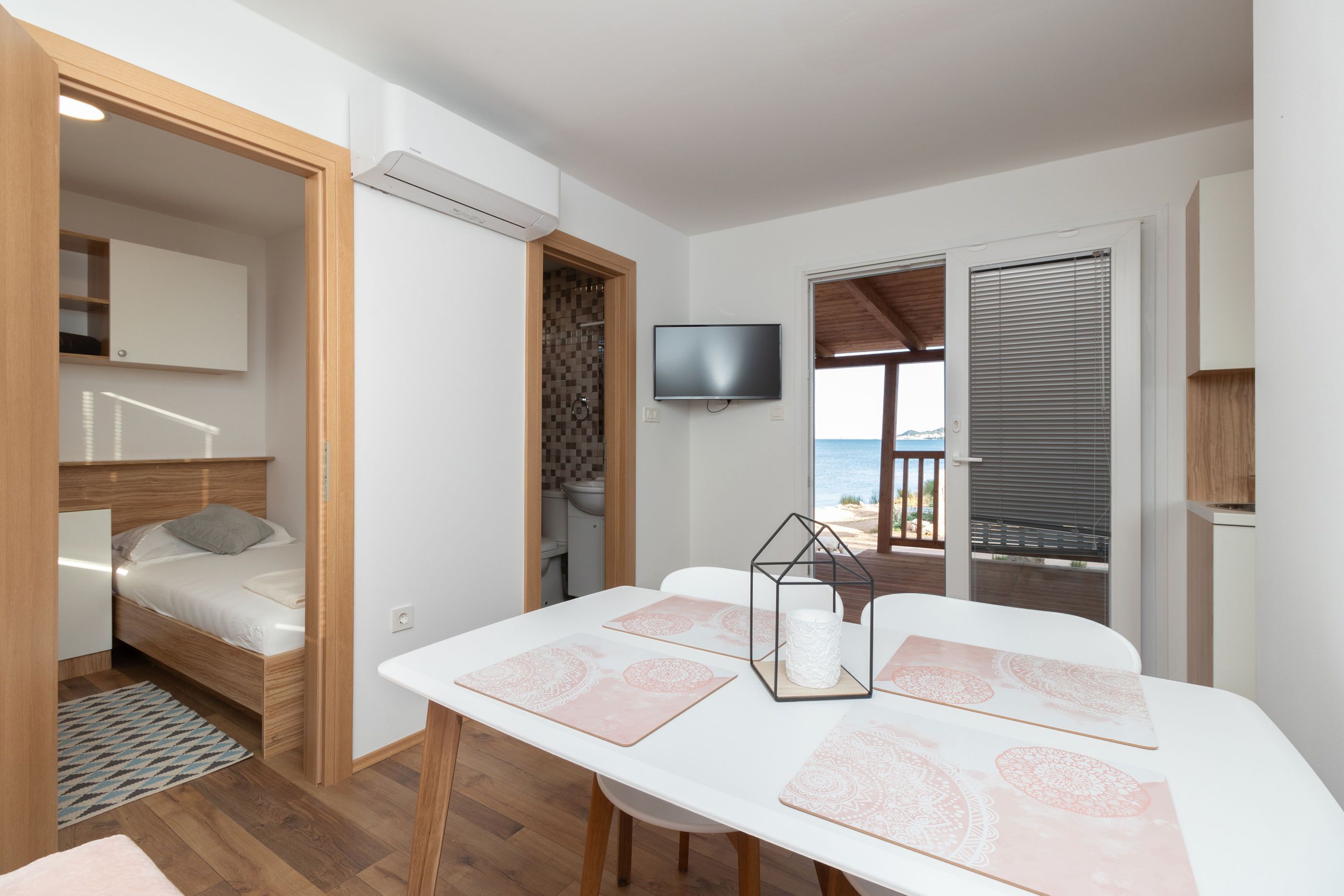 A compact room with a bed, a dining table with placemats, a television, and a view of the beach.
