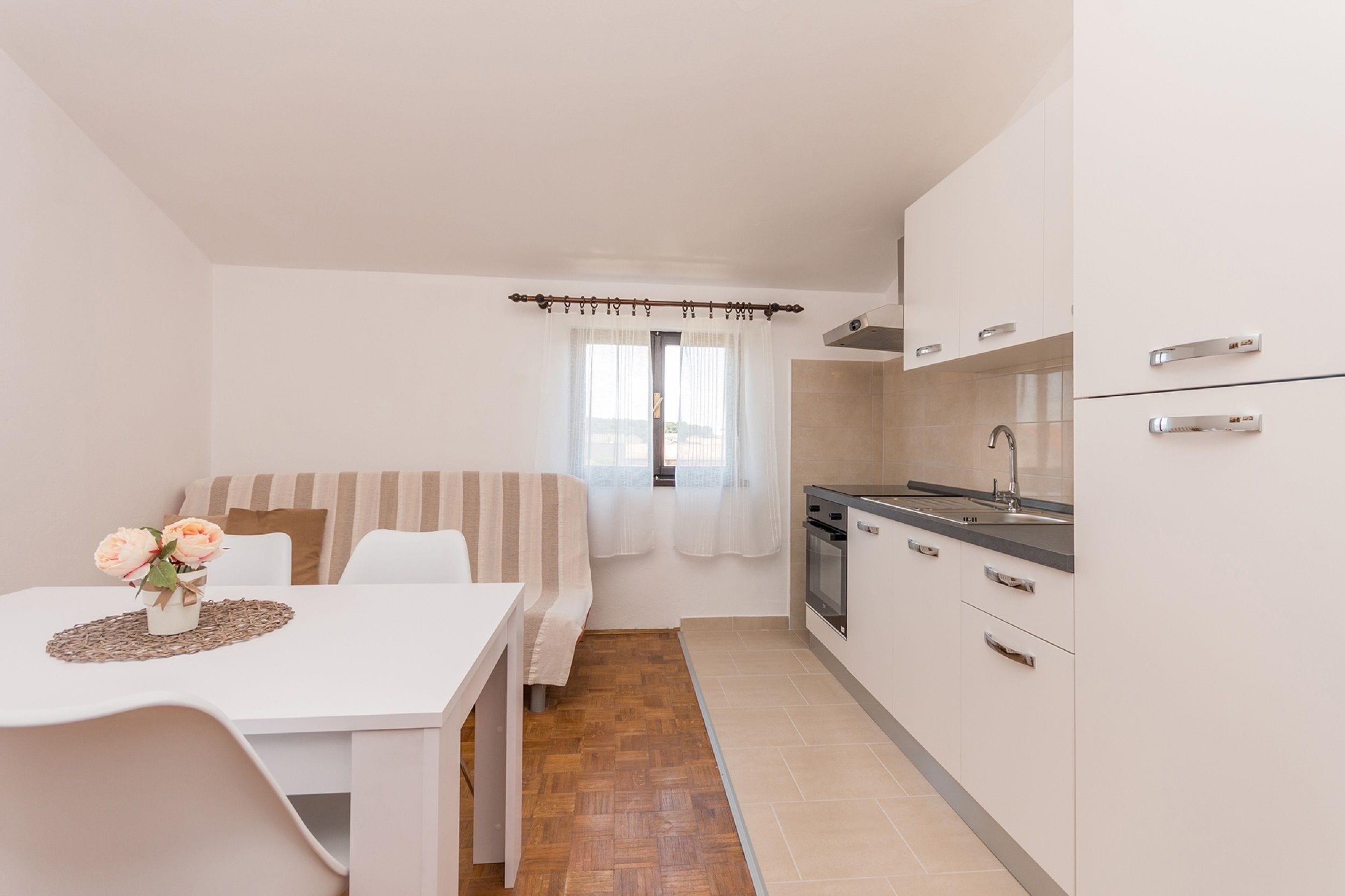 Een kleine keuken met witte kasten, een spoelbak en een kookplaat. Er is een eethoek met een witte tafel en een bloevas.
